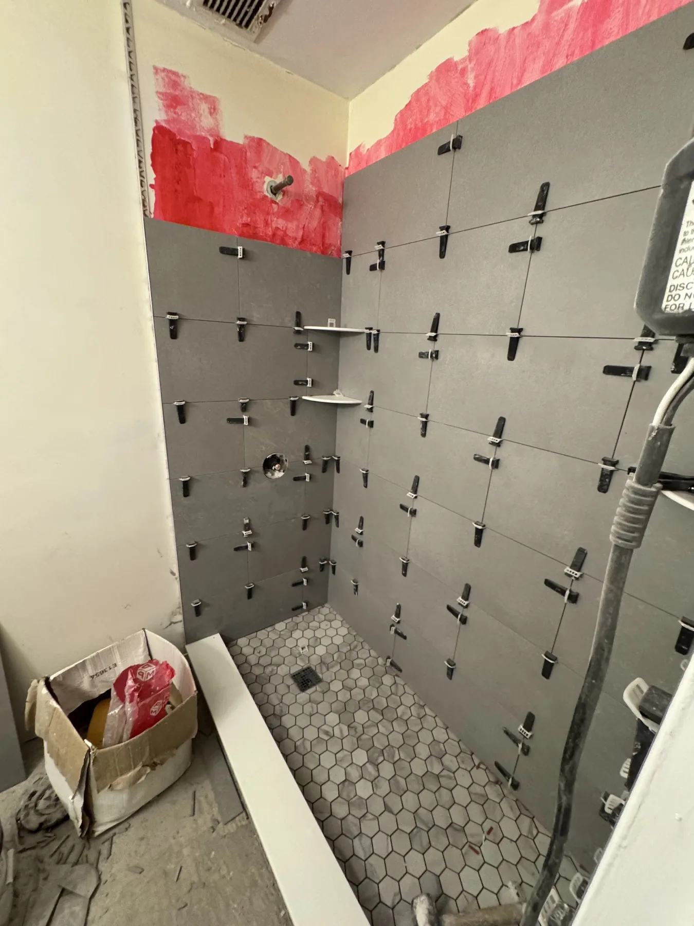 Gray shower wall tiles being installed with leveling clips above hexagonal floor tile